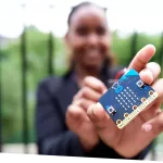 A child is standing outside in front of a metal fence with trees in the background, while holding a micro:bit controller up to the camera.