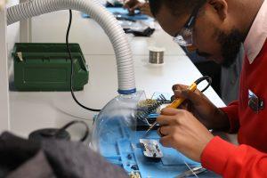 A delegate at the summer school leans over a white desk while holding a soldering iron in one hand. On the desk are a blue mat, tubing, and various printed circuit board components.