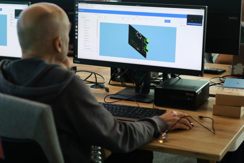 A delegate sits at a desk in a computer lab with his hand resting on a mouse, while looking toward a monitor where the 'Make Device' app is displayed.