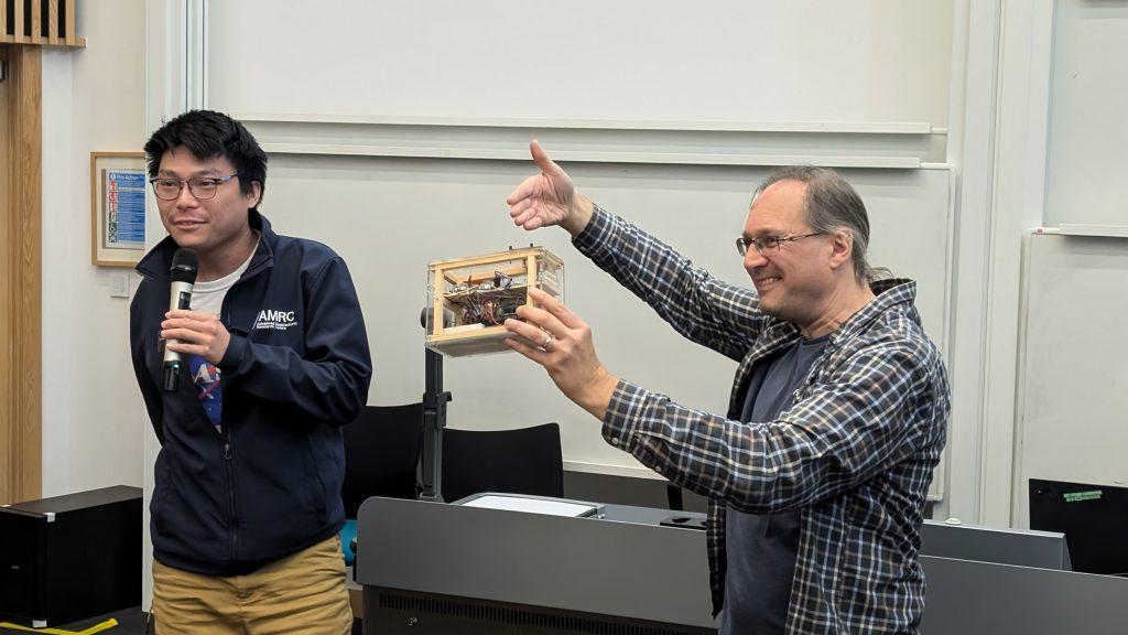 A delegate introduces himself at the front of a lecture theatre while holding a microphone. Beside him Professor Joe Finney holds a prototype on display to the audience while smiling. In the background a desk and whiteboards are visible.