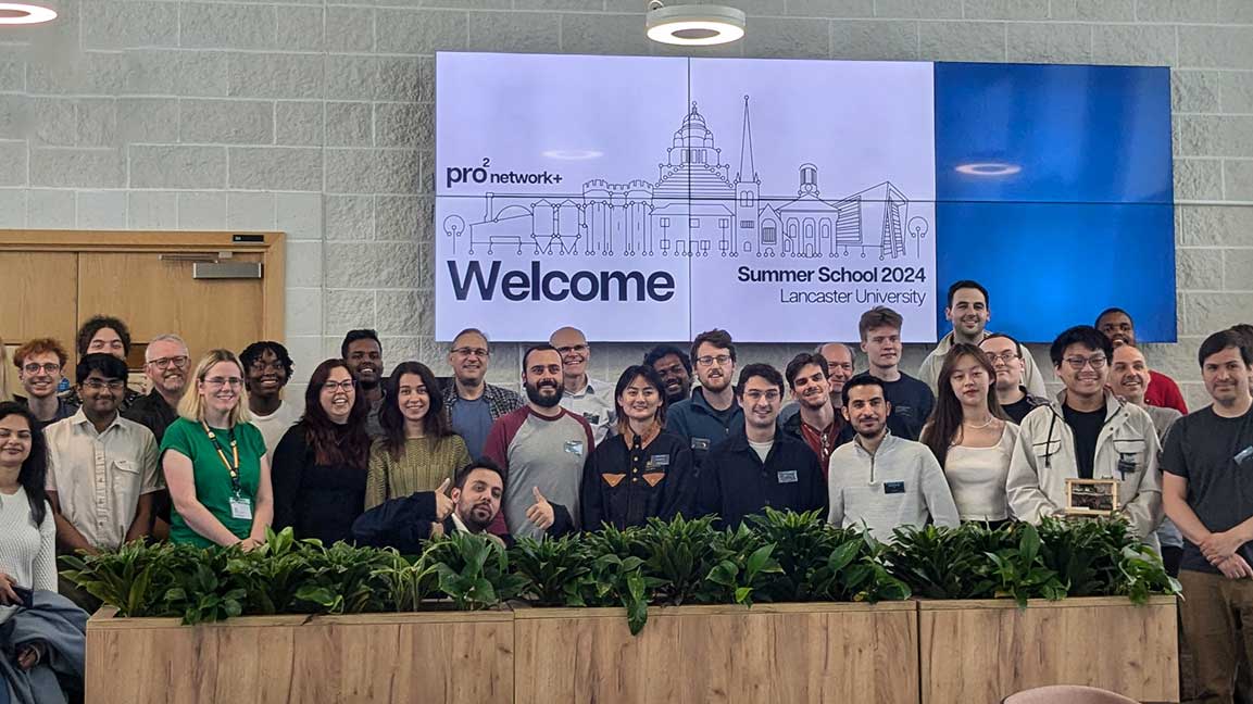 A photo of the 2024 pro2 summer school attendees standing against a white brick wall, with plants in the foreground and a large screen reading 'Welcome, Summer School 2024, Lancaster University' and the pro2 logo in the background.