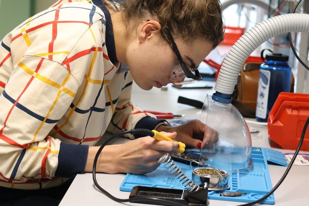 A summer school delegate leans over a desk in a lab while wearing safety goggles and holding a soldering iron.