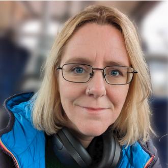 A colour photo of Lorraine Underwood, wearing glasses and a blue jacket against a blurry background. She's smiling and facing the camera.