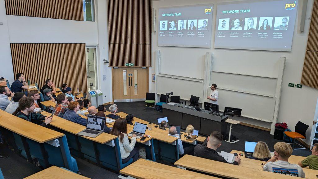 Professor Mike Fraser gives a talk in a crowded lecture theatre with two large screens above displaying details about the pro2 team.