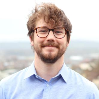 A photo in colour of Rory Clark, wearing glasses and a light blue shirt against a blurry outdoor background. He is facing the camera and smiling.