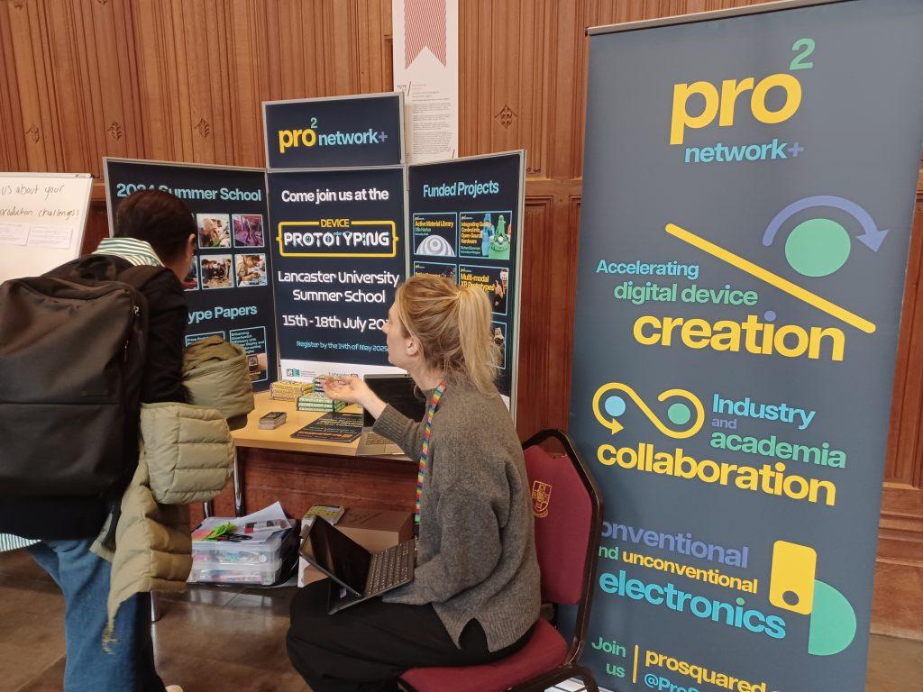 The pro2 network manager speaks to a Pre-CHI attendee who is viewing the network's table during the poster session. On the table are posters and chocolate bars, with a large banner to the right.
