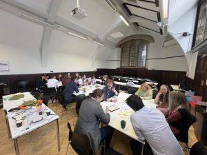 Attendees at a Pre-CHI workshop sit around large tables in groups, with paper, pens, and notebooks. One attendee is facing the camera and giving a thumbs up.