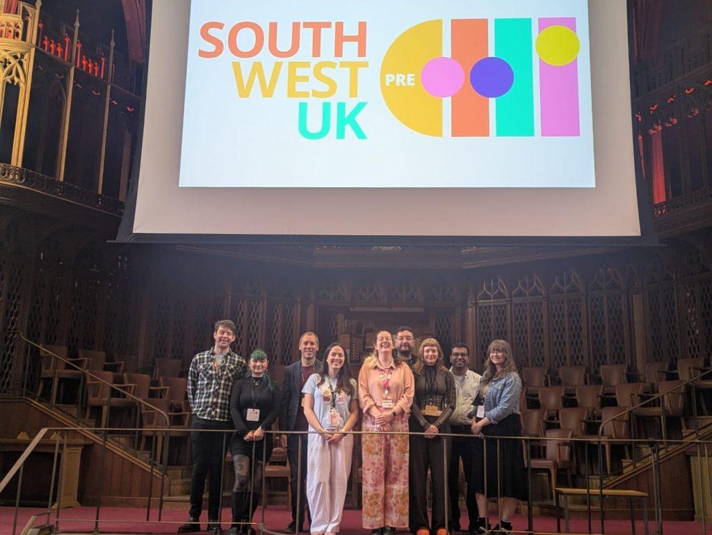Nine members of the South West UK Pre-CHI committee stand in the great hall at the University of Bristol underneath a large screen displaying the name of the event.