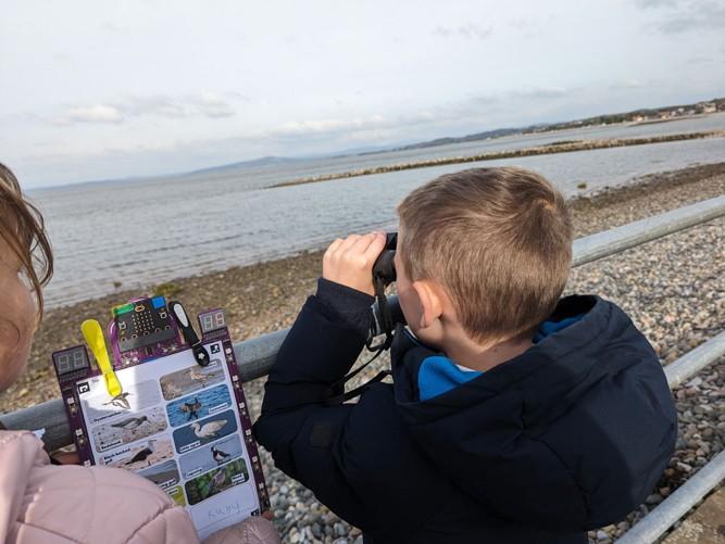 A photo shows two children at the beach, one of which is looking toward the ocean with binoculars while the other holds a clipboard device made with the micro:bit.