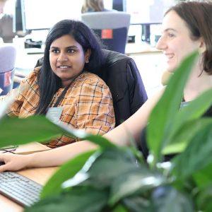Two summer school attendees sit together in a brightly-lit computer lab with leaves from a potted plant in the foreground.