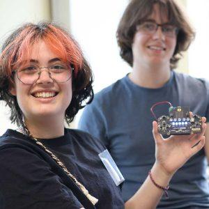 Two summer school attendees smile and hold up devices they've made to the camera. The devices are small and consist of a circuit board and wires.