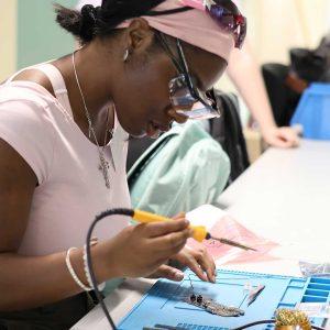 A summer school attendee holds a soldering iron in one hand while assembling a circuit board in a computer lab.