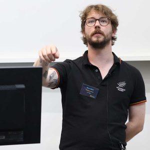 Rory Clark gives a talk at the front of a lecture hall, with one hand behind his back and the other resting on the edge of a computer monitor.
