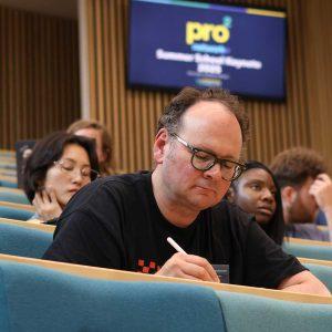 A summer school attendee sits in a crowded lecture theatre, looking down at a desk as he takes notes. A screen in the background displays the pro2 logo and name of the event.