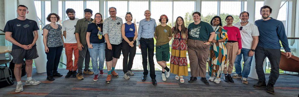 A row of fifteen people stand together in front of a large window at the 2025 summer school. They are all smiling and looking toward the camera.