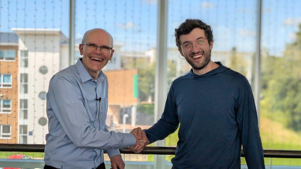 Steve Hodges shakes hands with Benjamin Seleb in front of a large window. Both are smiling widely and looking toward the camera. Outside blue skies and buildings can be seen.