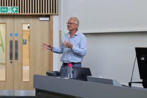 Simon Monk stands behind a desk with his hands spread as he delivers a talk in a lecture theatre.