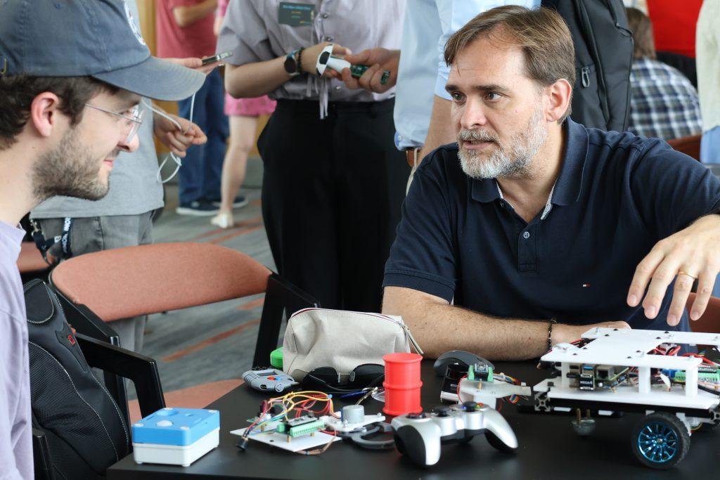 Two summer school attendees discuss electronic prototypes in a crowded room. On the table between them are a variety of electronic devices and components.