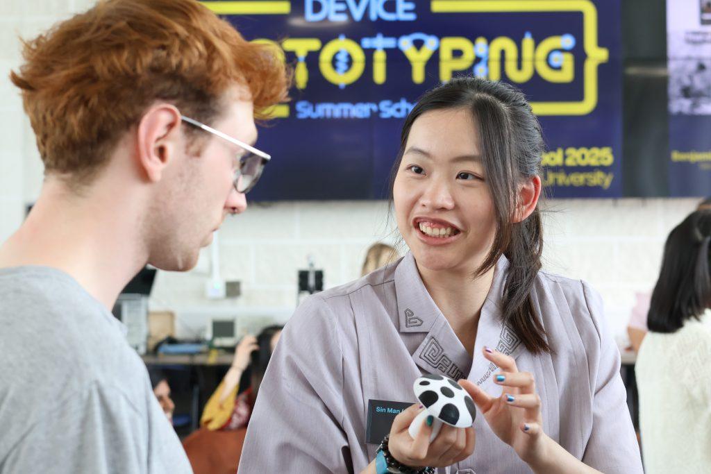 Two summer school attendees talk in a crowded room. One of them holds a small electronic device shaped like a mushroom. In the background a screen is displaying the event name.