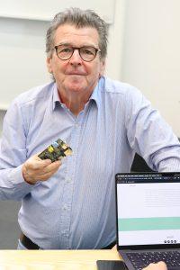 Pete Lomas smiles and faces the camera while standing in front of a desk and holding a Raspberry Pi circuit board.