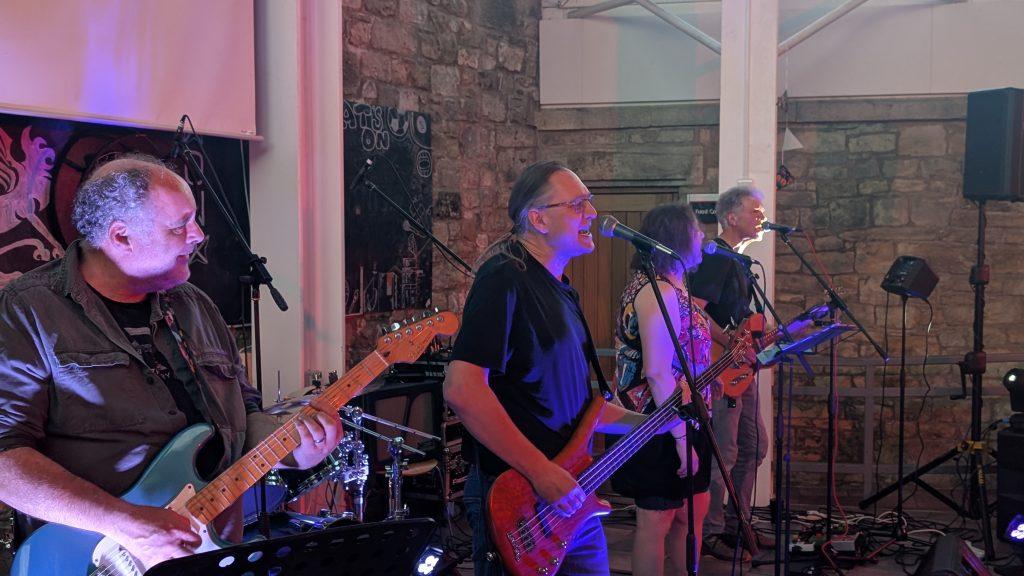 Four members of the band The Usuals can be seen singing and playing guitars in a university dining hall. Behind them a set of drums can be seen.