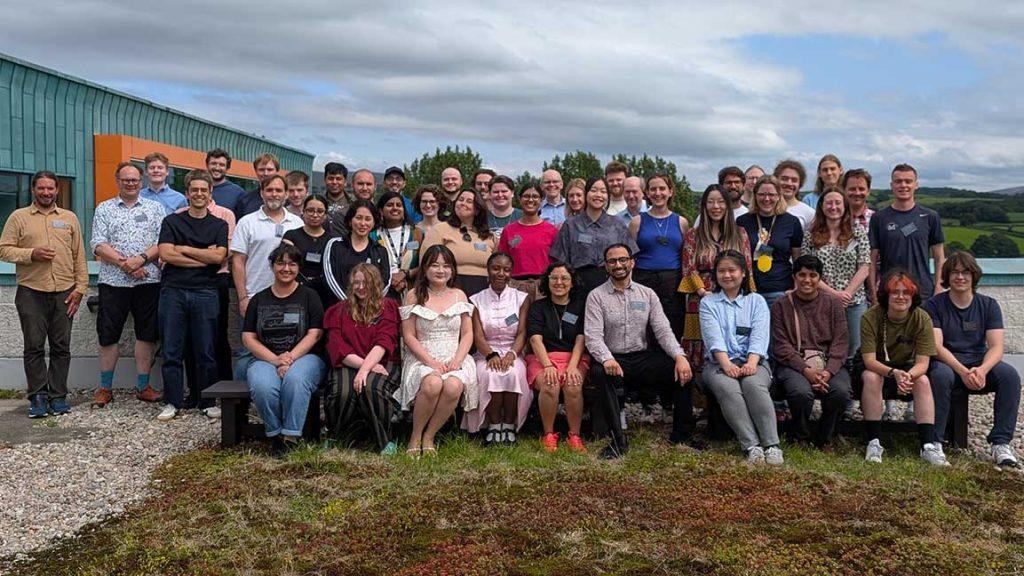 2025 summer school delegates sit and stand together outside on a cloudy day with a grassy lawn in front of them and a building to the left.