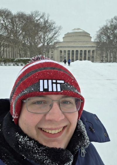 Researcher Joshua Brown smiles at the camera in a photo at MIT while wearing a red MIT wool hat and winter coat. In the background a snowy field, trees, and a building can be seen.